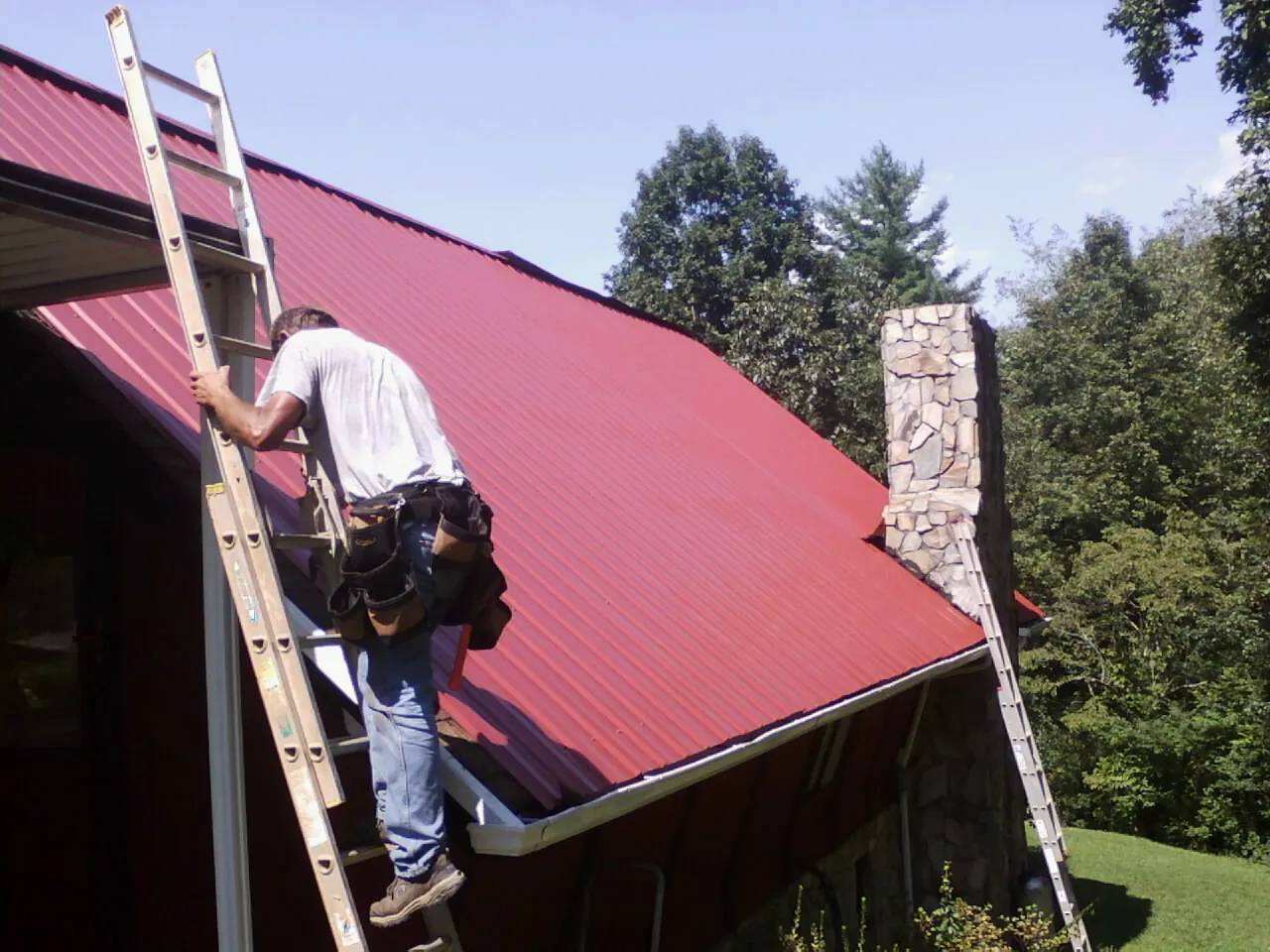 Roofer on a ladder beside a red metal roof and stone chimney — Soffit & Fascia Repair by Roofing Brothers in Columbus